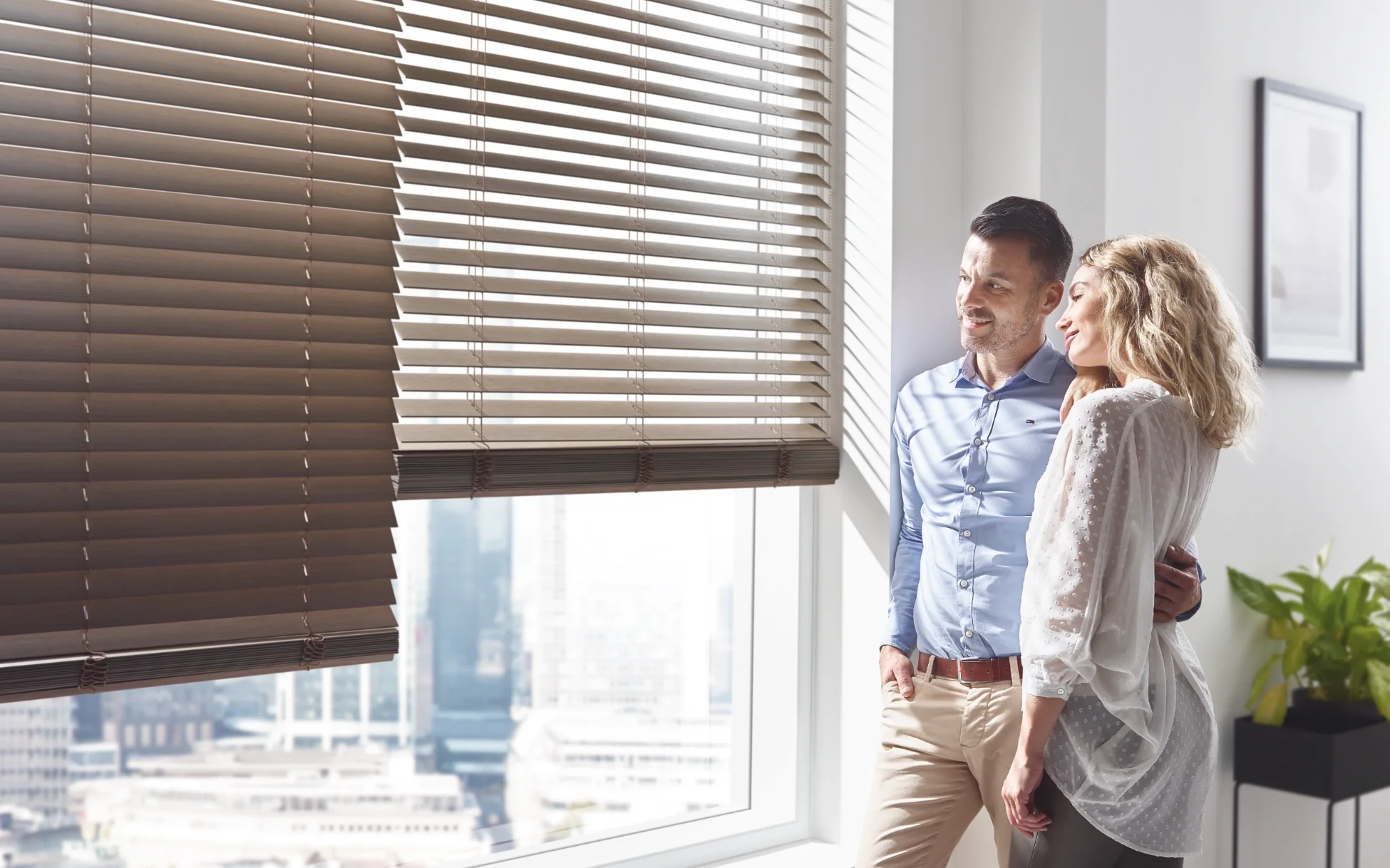 Jalousie aus Holz vor einem Großen Fenster mit einem Paar, dass aus dem Fenster schaut.